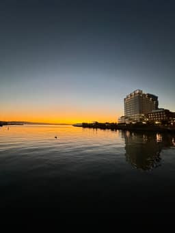 A modern waterfront building complex reflects in calm waters during sunset, with warm golden light illuminating the horizon and a small bird visible on the water's surface.