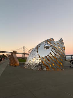 A large metallic fish sculpture with reflective scales and a circular eye sits on a waterfront plaza at sunset, with the Bay Bridge visible in the background.